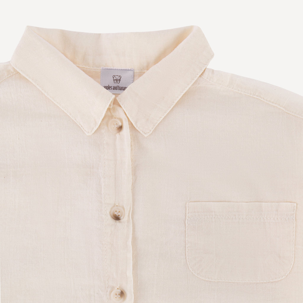 The image depicts a close-up view of a beige long-sleeve button-up shirt. It showcases the collar, a single chest pocket, and a row of buttons. The fabric appears to be lightweight and breathable. The label on the shirt reads "apples and bananas."