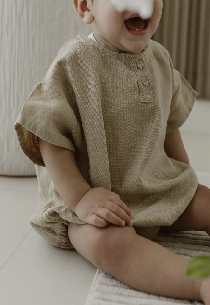 A baby wearing a light caramel gray romper from Apples and Bananas sits on a ground surface. The background, featuring a cozy living room setting , enhances the warm and inviting atmosphere. This adorable scene perfectly captures the essence of comfort and simplicity, making it an ideal moment for the Apples and Bananas brand.