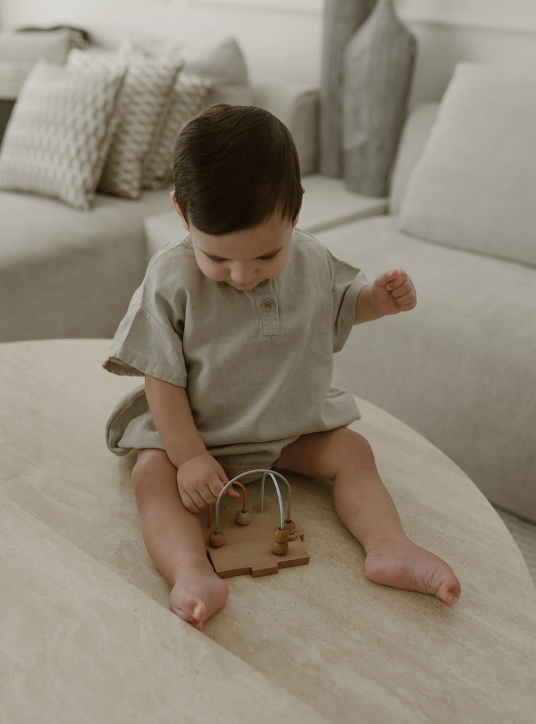 A baby wearing a beige romper from Apples and Bananas is sitting on a wooden surface, playing with a wooden toy. The background features a cozy living room setting with neutral-colored furniture and pillows.