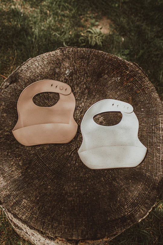 Two bibs from Apples and Bananas, one beige and one off-white, placed on a wooden tree stump with a background of grass.