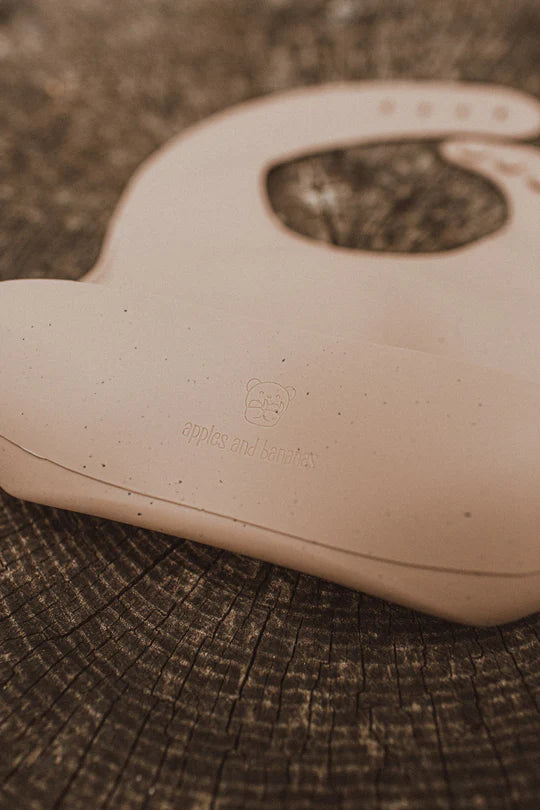 A close-up of a beige bib from Apples and Bananas, featuring their logo, placed on a wooden tree stump background.