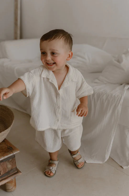 A happy toddler in a cream-colored Apples and Bananas outfit stands indoors beside a wooden stool.