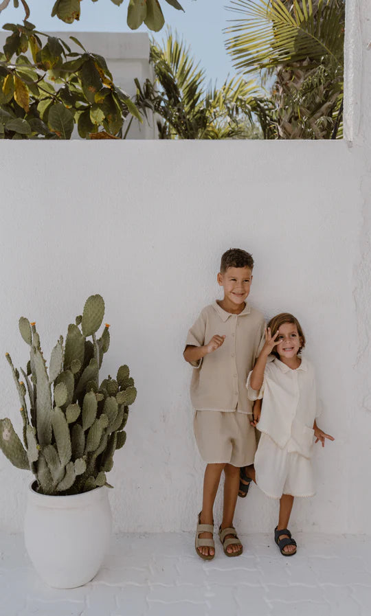 Two young children smiling and standing next to a white wall with a cactus plant in a pot, both wearing beige ravi set of cream-colored outfits by apples and bananas.