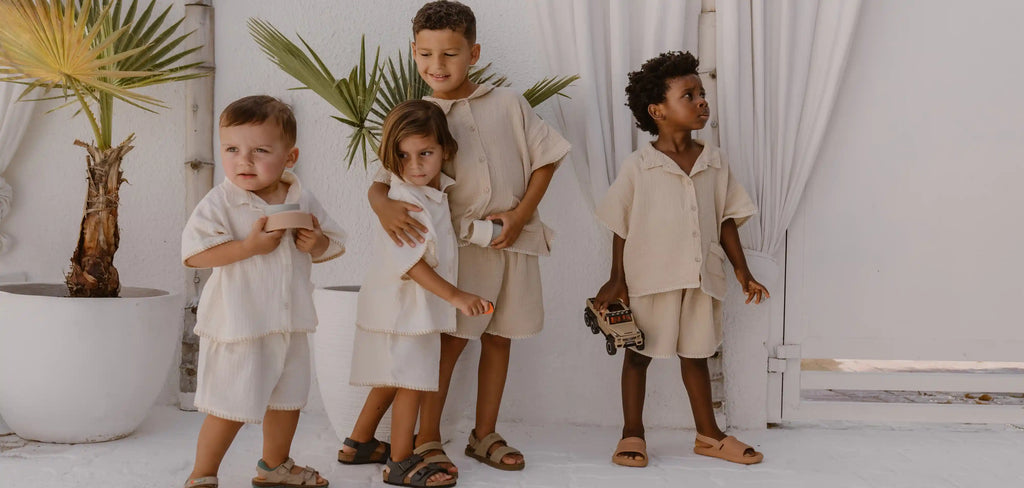 Four children in summer outfits, likely from the Apples and Bananas brand, stand against a white background with palm plants. The child on the left holds a bowl and wears a white shirt with matching shorts. The next two children, both in beige outfits, stand close together, with the taller one smiling and the shorter one having shoulder-length hair. The child on the right holds a toy car and also wears beige. All the children are wearing sandals.